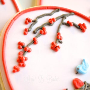 May include: A close-up of a heart-shaped cookie decorated with red and gray icing. The icing depicts a branch with red berries and two blue birds. The cookie is surrounded by a red border.