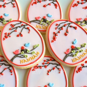 May include: A set of six decorated sugar cookies with a pink background, a brown branch with green leaves, red berries, and two blue and red birds. The cookies are decorated with the name "HANNAH" in yellow icing.
