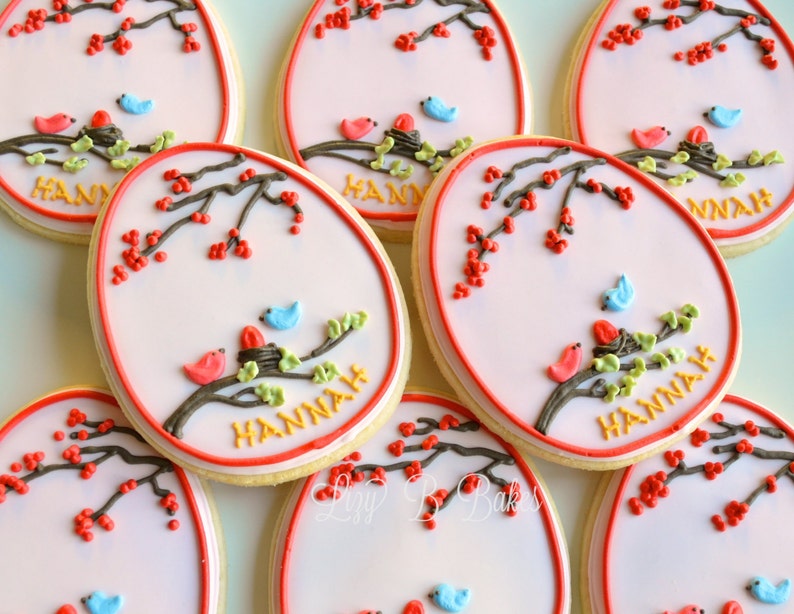 May include: A set of six decorated sugar cookies with a pink background, a brown branch with green leaves, red berries, and two blue and red birds. The cookies are decorated with the name "HANNAH" in yellow icing.