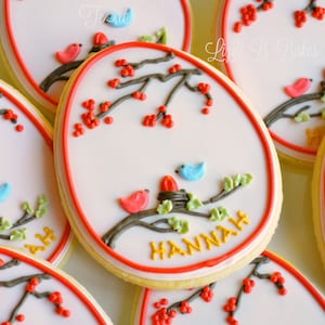May include: A set of decorated sugar cookies with a red, white, and blue color scheme. The cookies feature a branch with red berries, green leaves, and small birds. One cookie has the name "HANNAH" written in yellow icing.
