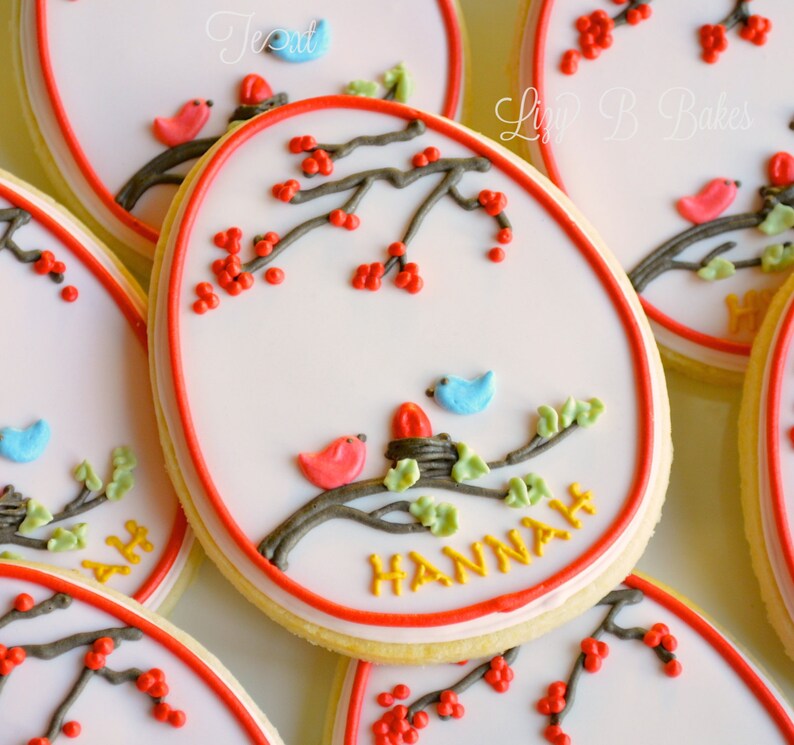 May include: A set of decorated sugar cookies with a red, white, and blue color scheme. The cookies feature a branch with red berries, green leaves, and small birds. One cookie has the name "HANNAH" written in yellow icing.