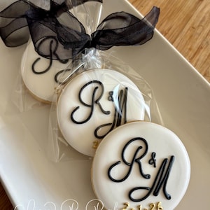 May include: Three round cookies with white icing, black initials, and gold numbers are packaged in clear cellophane and tied with a black ribbon. The cookies are on a white rectangular plate.
