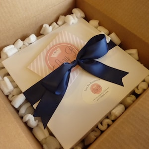 May include: A square white box inside a cardboard shipping container filled with white foam packing peanuts. The box is tied with a dark navy blue satin ribbon and features a circular pink logo sticker with a crown and the text Lily B.