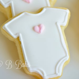 May include: A sugar cookie shaped like a baby onesie, decorated with smooth white icing, a raised white piped border, and a small light pink heart in the center. The text Lizy B Bakes is visible in the lower left corner of the image.