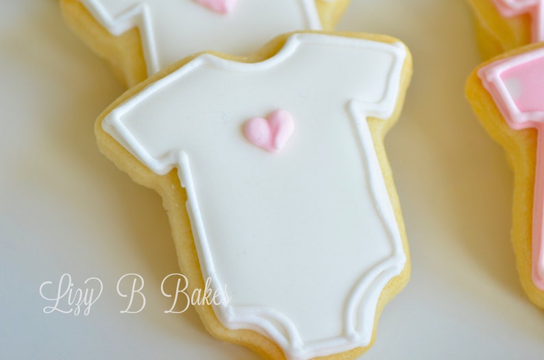 May include: A sugar cookie shaped like a baby onesie, decorated with smooth white icing, a raised white piped border, and a small light pink heart in the center. The text Lizy B Bakes is visible in the lower left corner of the image.