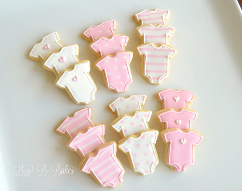 May include: A collection of baby onesie-shaped sugar cookies decorated with royal icing. The cookies feature various designs including solid pink, solid white, pink and white stripes, and pink and white polka dots, some accented with small hearts.