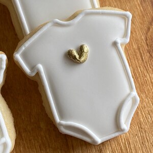 以下が含まれることがあります： 白い砂糖菓子は、白いアイシングと金色のハートで飾られており、ベビーロンパースの形をしています。