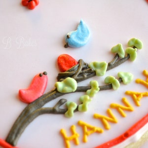 May include: A white and red decorated cookie with a brown branch with green leaves and red and blue birds. The cookie is decorated with the name "HANNAH" in yellow icing.