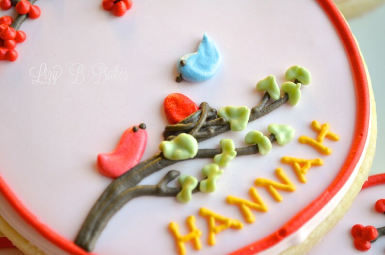 May include: A white and red decorated cookie with a brown branch with green leaves and red and blue birds. The cookie is decorated with the name "HANNAH" in yellow icing.
