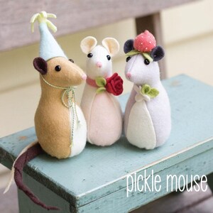 May include: Three handcrafted felt mice, each with distinct adornments. One sports a party hat, another a red rose, and the third a strawberry. The mice are displayed on a pale blue surface, with the text "pickle mouse" visible.