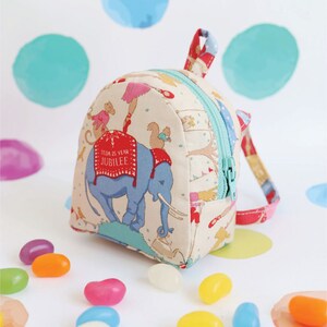 May include: A small, cream-coloured backpack with a red and blue zipper. The backpack is decorated with a circus theme, including an elephant, a monkey, and a squirrel. The text "100th 25 Year Jubilee" is printed on the backpack.