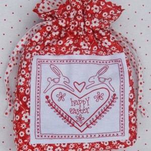 May include: Red and white fabric drawstring bag with a white embroidered panel featuring two bunnies hopping towards a heart with the text "happy easter" inside the heart.