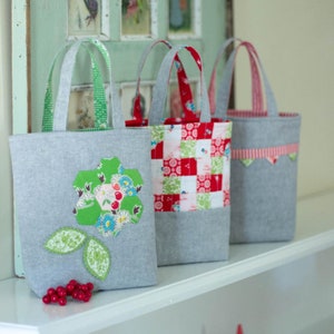 May include: Three grey fabric tote bags with different patterned fabric accents. The first bag has a green flower appliqué, the second has a red and white checked pattern, and the third has a red and white scalloped trim.