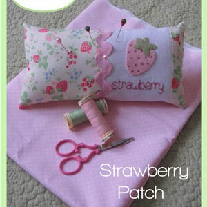 May include: A handmade strawberry patch pincushion with a floral print pillow and a strawberry-shaped design. The image includes pink fabric, thread spools, and pink scissors. The pincushion is decorated with pins.