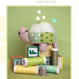 May include: A turtle-shaped pincushion with a patchwork shell, felt head and legs, and pins. The image also shows spools of thread, wooden blocks, and a thimble. The text reads "Tiny Tim Pincushion" and "Creative Card".