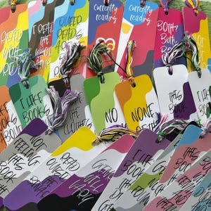 May include: A collection of colorful bookmarks with various text designs and tassel accents. The bookmarks feature phrases like "I like books," "Queer Reads," and "Currently Reading." Each bookmark has a unique color scheme, including orange, green, purple, and yellow.