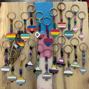 May include: A collection of twenty-four metal keychains featuring various pride flag designs in heart, diamond, and spade shapes. Each charm is attached to a circular metal ring by a short, multi-coloured chain made of small, interlocking metal jump rings.
