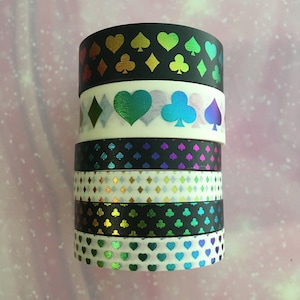 May include: A stack of six washi tapes with different designs. The top tape has a black background with iridescent rainbow coloured playing card suits. The second tape has a white background with iridescent rainbow coloured playing card suits. The third tape has a black background with iridescent rainbow coloured diamonds. The fourth tape has a white background with iridescent rainbow coloured diamonds. The fifth tape has a black background with iridescent rainbow coloured playing card suits. The bottom tape has a white background with iridescent rainbow coloured hearts.