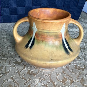 May include: A ceramic vessel with two handles, featuring a tan base color with a visible crackle glaze finish. The body is decorated with horizontal bands of muted green and brown, accented by two vertical white and black abstract leaf-like motifs.