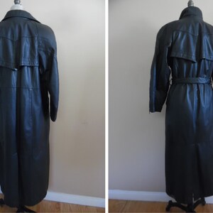 Op de afbeelding: Een lange zwarte leren trenchcoat met een riem en een kraag. De jas heeft een enkele rij knopen en een split in de rug.
