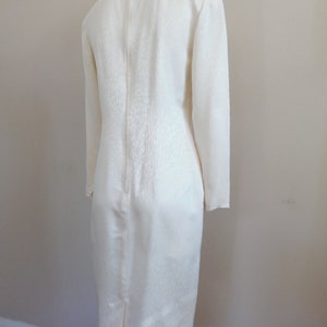 Pode incluir: Vestido cor marfim com mangas compridas e bainha at&eacute; ao joelho. O vestido apresenta um tecido texturizado e uma subtil guarni&ccedil;&atilde;o dourada &agrave; volta do decote. Um fecho corre pela parte de tr&aacute;s do vestido.