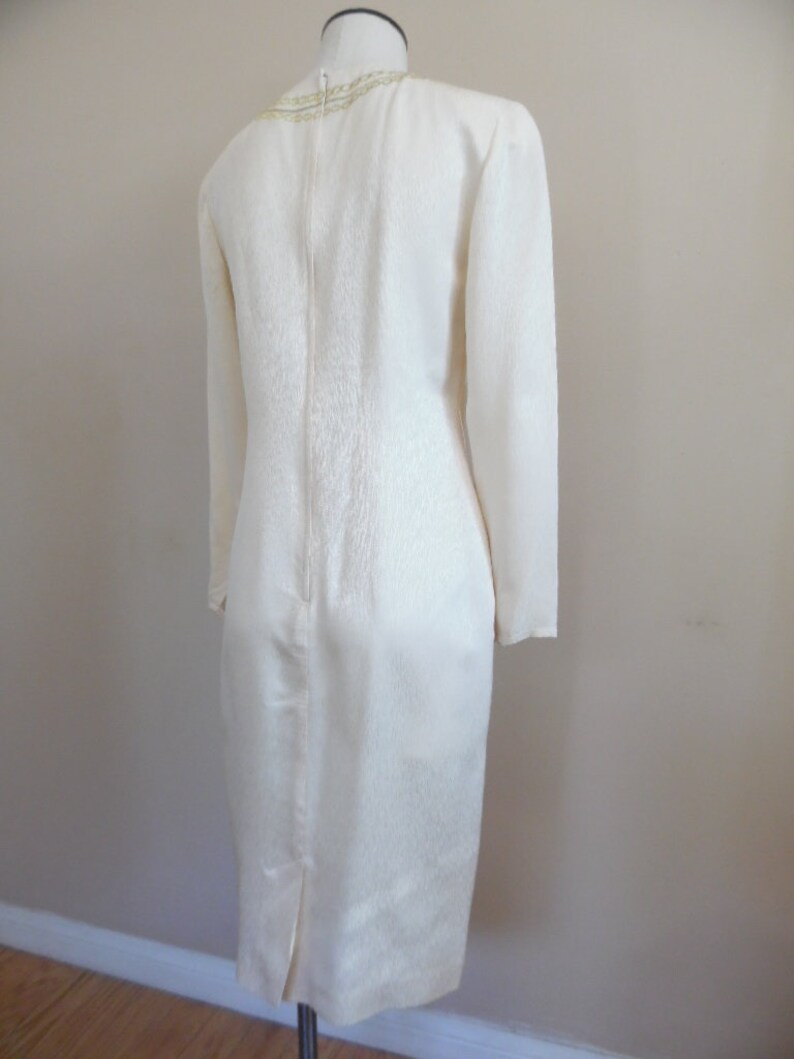 Pode incluir: Vestido cor marfim com mangas compridas e bainha at&eacute; ao joelho. O vestido apresenta um tecido texturizado e uma subtil guarni&ccedil;&atilde;o dourada &agrave; volta do decote. Um fecho corre pela parte de tr&aacute;s do vestido.