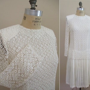 May include: White lace dress with a pleated skirt. The dress has long sleeves and a round neckline. The lace is a delicate pattern of small circles and flowers.