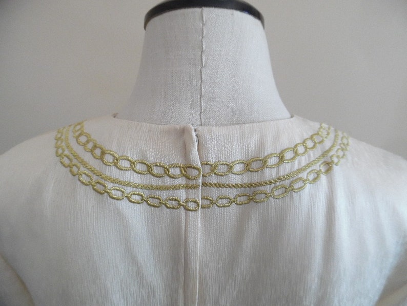 Pode incluir: Vestu&aacute;rio creme com bordado dourado de elos de corrente e corda ao redor do decote. O bordado &eacute; disposto em tr&ecirc;s fileiras, criando um efeito de gola decorativa. O tecido tem uma textura sutil.