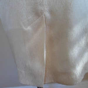 Pode incluir: Close-up de uma pe&ccedil;a de roupa creme com uma superf&iacute;cie texturizada. O tecido tem um brilho subtil e uma fenda &eacute; vis&iacute;vel na bainha inferior. A pe&ccedil;a de roupa parece ser uma saia ou um vestido.