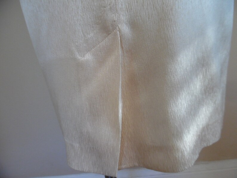 Pode incluir: Close-up de uma pe&ccedil;a de roupa creme com uma superf&iacute;cie texturizada. O tecido tem um brilho subtil e uma fenda &eacute; vis&iacute;vel na bainha inferior. A pe&ccedil;a de roupa parece ser uma saia ou um vestido.