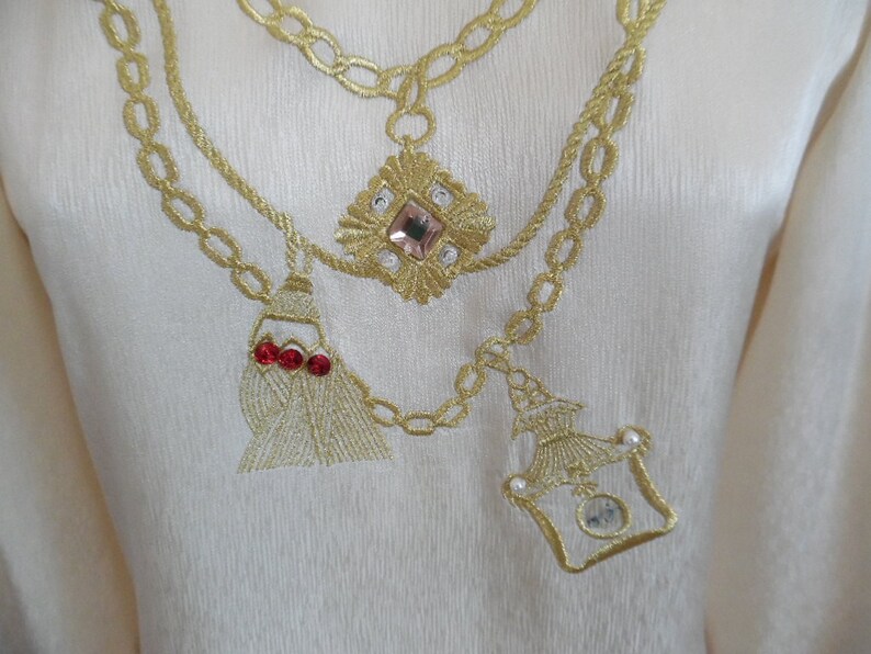 Pode incluir: Tecido creme com bordado dourado. O bordado apresenta um design de corrente com uma pedra preciosa rosa, pedras preciosas vermelhas e um frasco de perfume decorativo. O tecido tem uma textura sutil.