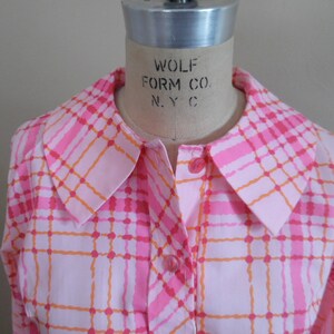 May include: A pink and orange plaid shirt with a large collar. The shirt has a button-up front and long sleeves. The label on the collar reads "Wolf Form Co. N.Y.C."