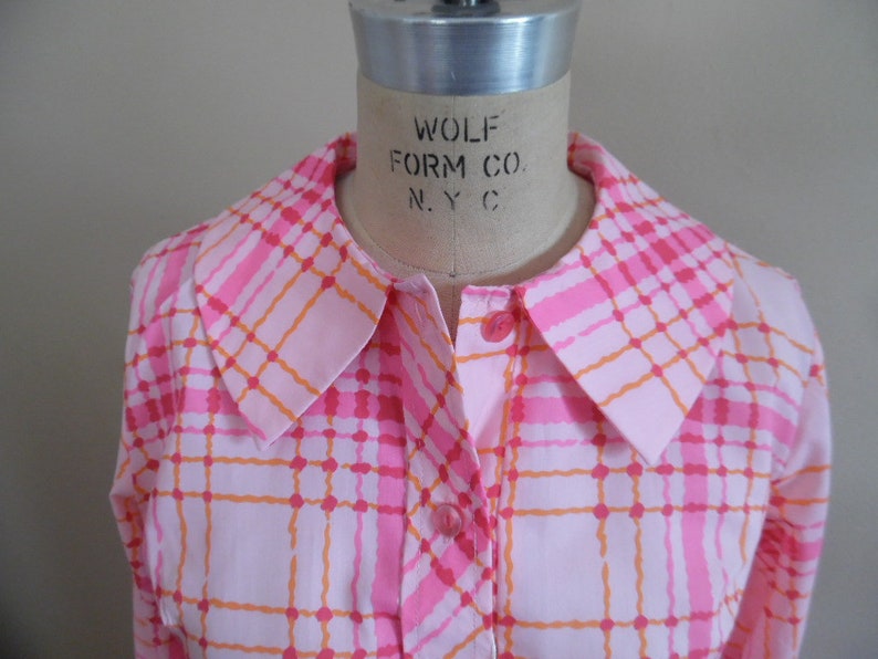May include: A pink and orange plaid shirt with a large collar. The shirt has a button-up front and long sleeves. The label on the collar reads "Wolf Form Co. N.Y.C."