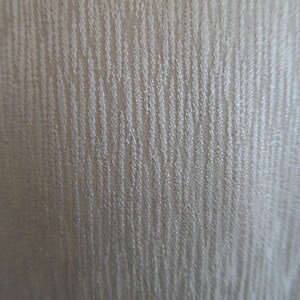 Pode incluir: Close-up de um tecido texturizado com um padr&atilde;o vertical sutil. O tecido tem uma cor clara e neutra, com um brilho ligeiramente iridescente. A textura parece ser tecida ou em relevo, criando uma profundidade visual.