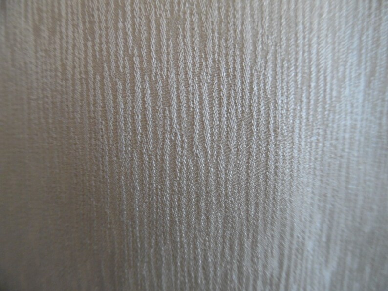 Pode incluir: Close-up de um tecido texturizado com um padr&atilde;o vertical sutil. O tecido tem uma cor clara e neutra, com um brilho ligeiramente iridescente. A textura parece ser tecida ou em relevo, criando uma profundidade visual.