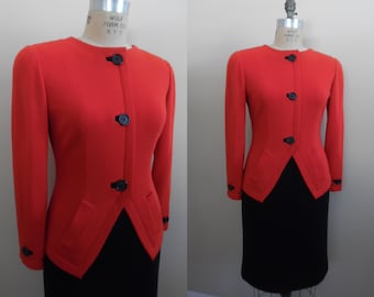 May include: A two-piece suit featuring a bright red jacket and a black skirt. The jacket has a round neckline, three dark buttons, and two front pockets. The skirt is a knee-length pencil skirt. The suit is displayed on a mannequin.