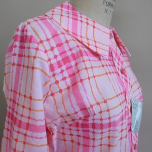 May include: A pink and orange plaid button-up shirt with a collared neckline. The shirt has a pocket on the left side and a tag that says "Size XS".