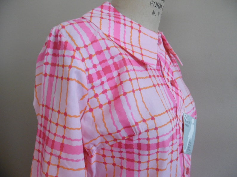 May include: A pink and orange plaid button-up shirt with a collared neckline. The shirt has a pocket on the left side and a tag that says "Size XS".