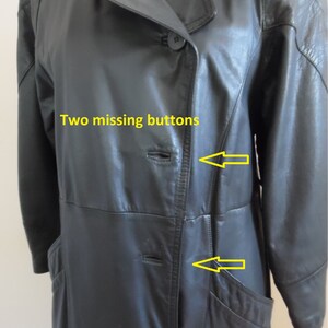 Op de afbeelding: Een zwarte leren trenchcoat met twee ontbrekende knopen. De knopen bevinden zich aan de voorkant van de jas, in de buurt van de zakken.