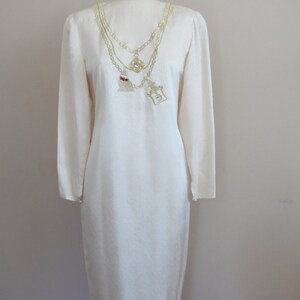 Pode incluir: Vestido creme de manga comprida com um design de corrente dourada decorativa. A corrente apresenta um pingente com uma borla vermelha e um pingente quadrado com uma pedra transparente. O vestido &eacute; exibido em um manequim.