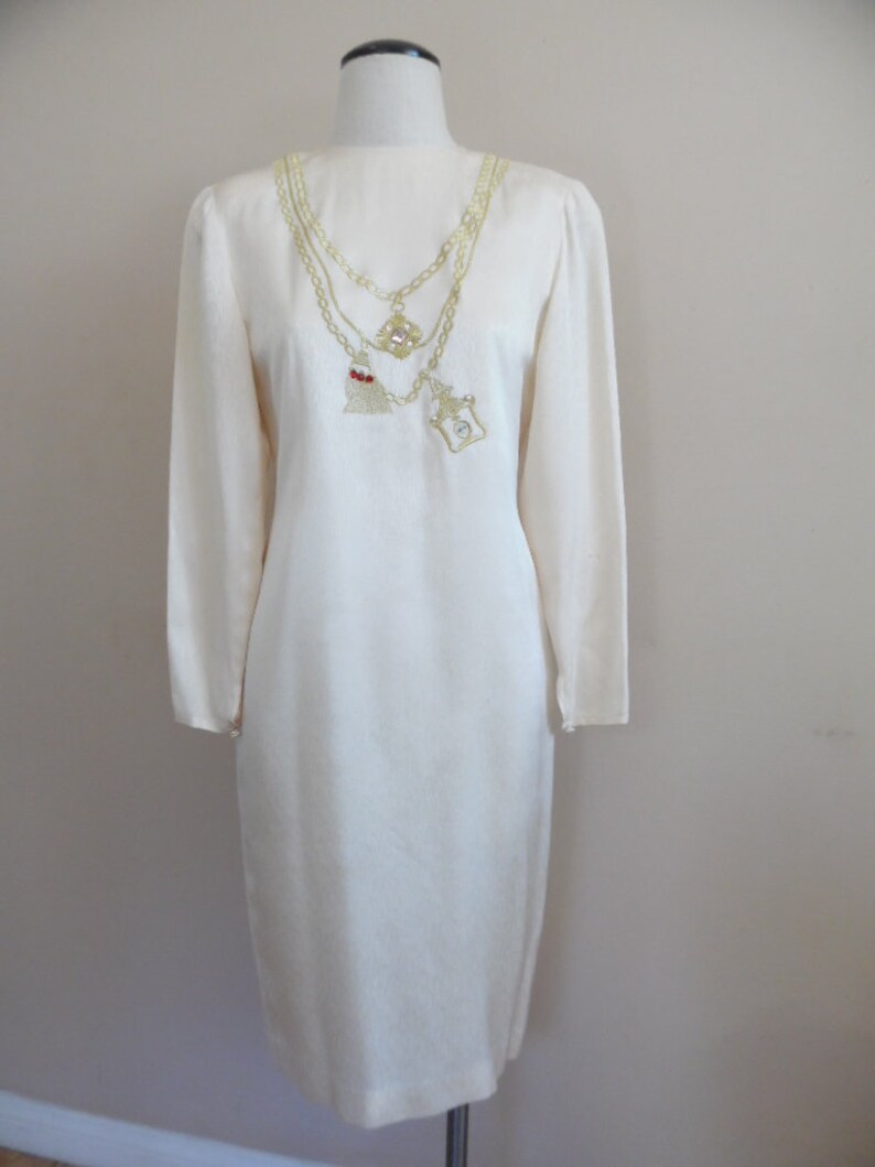 Pode incluir: Vestido creme de manga comprida com um design de corrente dourada decorativa. A corrente apresenta um pingente com uma borla vermelha e um pingente quadrado com uma pedra transparente. O vestido &eacute; exibido em um manequim.