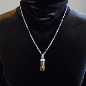Pode incluir: Um colar de corrente de prata com um pequeno pingente de frasco de vidro transparente cheio de pedras coloridas e iridescentes.