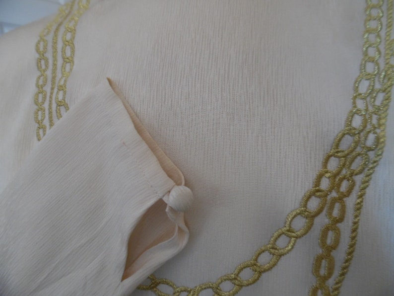Pode incluir: Vestu&aacute;rio creme de manga comprida com acabamento dourado em forma de corrente ao redor do decote e da manga. A manga tem um fecho de bot&atilde;o. O tecido tem um aspeto texturizado.