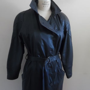 Op de afbeelding: Een lange zwarte leren trenchcoat met een riem en een dubbele knoopsluiting. De jas heeft een kraag en zakken. Het label op de jas luidt "Wolf Form Co. N.Y.C."
