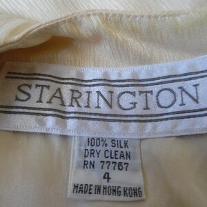 Pode incluir: Close-up de uma pe&ccedil;a de roupa creme com uma etiqueta que diz "STARINGTON" em texto cinza. A etiqueta tamb&eacute;m indica que a pe&ccedil;a &eacute; 100% seda, apenas limpeza a seco, tamanho 4 e fabricada em Hong Kong.