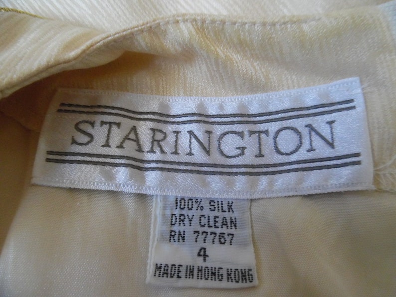 Pode incluir: Close-up de uma pe&ccedil;a de roupa creme com uma etiqueta que diz "STARINGTON" em texto cinza. A etiqueta tamb&eacute;m indica que a pe&ccedil;a &eacute; 100% seda, apenas limpeza a seco, tamanho 4 e fabricada em Hong Kong.