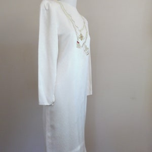 Pode incluir: Vestido marfim de manga comprida com decote redondo. O vestido &eacute; exibido em um manequim. Um colar em camadas com pingentes pende na frente. O vestido tem um design simples e elegante.