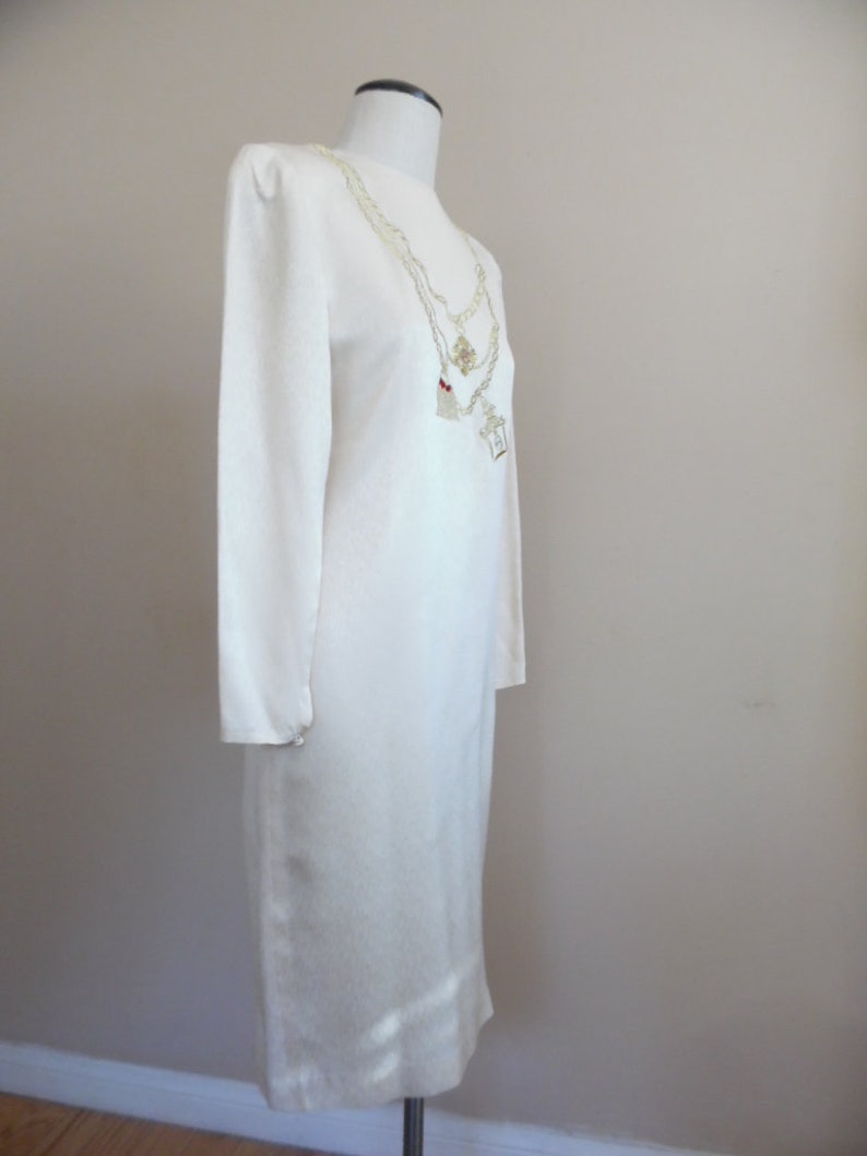 Pode incluir: Vestido marfim de manga comprida com decote redondo. O vestido &eacute; exibido em um manequim. Um colar em camadas com pingentes pende na frente. O vestido tem um design simples e elegante.