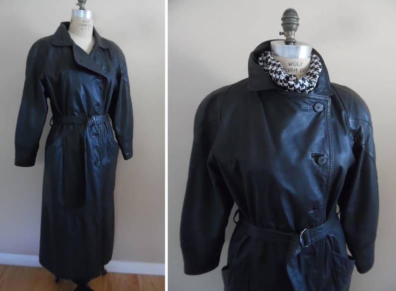 Op de afbeelding: Een lange zwarte leren trenchcoat met een riem en knoopsluiting. De jas heeft een kraag en zakken.