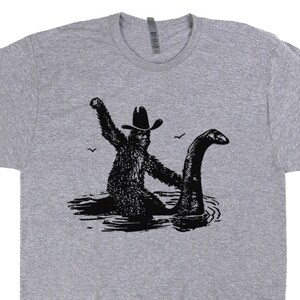 May include: A heather grey t-shirt with a black and white graphic of a creature from the Loch Ness monster riding a cowboy on a lake.